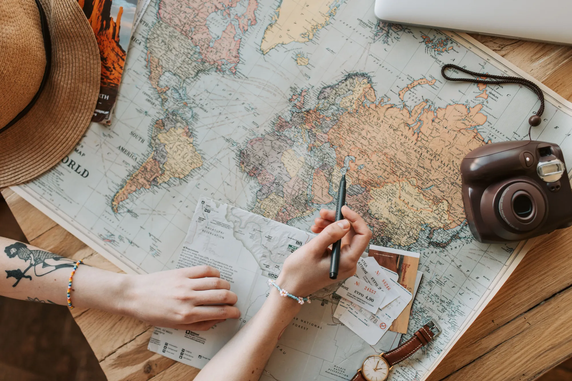 A traveler planning a route on a world map, surrounded by a hat, camera, and travel tickets