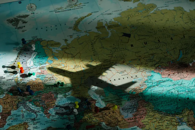 A world map marked with pins, with the shadow of an airplane flying overhead